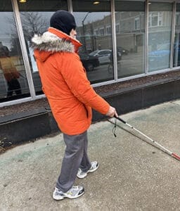 Man walking with white cane on a sidewalk. He is wearing an orange coat and a black winter hat