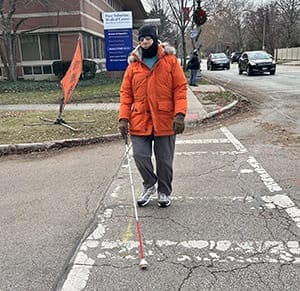 Man crossing a street in a crosswalk with a white cane. He is wearing an orange coat and a black winter hat.