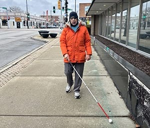 Man walking toward camera on a sidewalk with a white cane. He's wearing an orange coat and black winter hat.