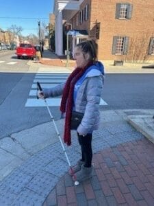 A woman standing on a street corner in winter gear holding a white cane. 