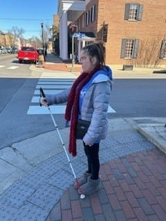 A woman standing on a street corner in winter gear holding a white cane.