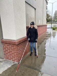 A woman walking on a wet sidewalk with a white cane and stabilizing cane.