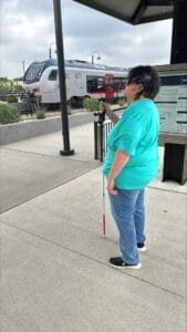 Judy holding her white cane at a train station waiting for a train to arrive.