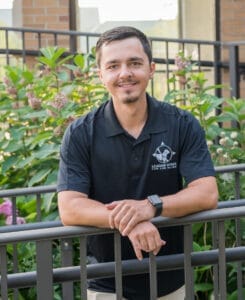 A man leans against a black fence. He is wearing a sporty black polo with the Leader Dog for the Blind logo on it. He has dark hair and a mustache and goatee.