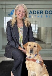 A woman, Missy Aupperle, kneels down next to a yellow Labrador Leader Dog in harness. The Leader Dogs for the Blind logo is seen on the glass doors behind her.