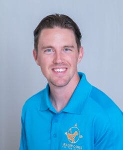 A man wearing a light blue polo with the yellow Leader Dogs for the Blind logo on it, sits infront of a light background and looks to the camera smiling.