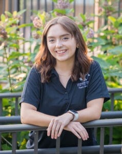 A young woman with shoulder length brown hair leans against a black fence. She is wearing a black sporty polo with the Leader Dogs for the Blind logo on it.
