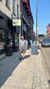 A man walking along a downtown area sidewalk using a white cane.