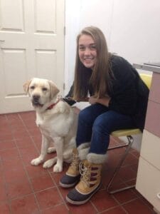 A young woman sits on a chair, she is wearing a black long sleeve shirt and boots. Next to her is yellow Labrador retriever.
