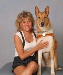 A woman with light brown medium lenght hair sits against a light colored background. Sitting next to her is a yellow and white Leader Dog in harness.