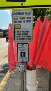 Sign instructing pedestrians to use crosswalk flags and push a button for a crosswalk warning device located at a street crossing. Red flags are provided to alert traffic before entering the crosswalk. Safety instructions urge caution.