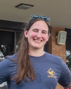 A woman with long, light brown hair smiles at the camera. She is wearing a dark blue t-shirt with the Leader Dogs for the Blind logo printed in orange over her left shoulder.