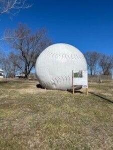 A large, round structure designed to look like a baseball, with stitches details, placed in a grassy field under a clear blue sky.