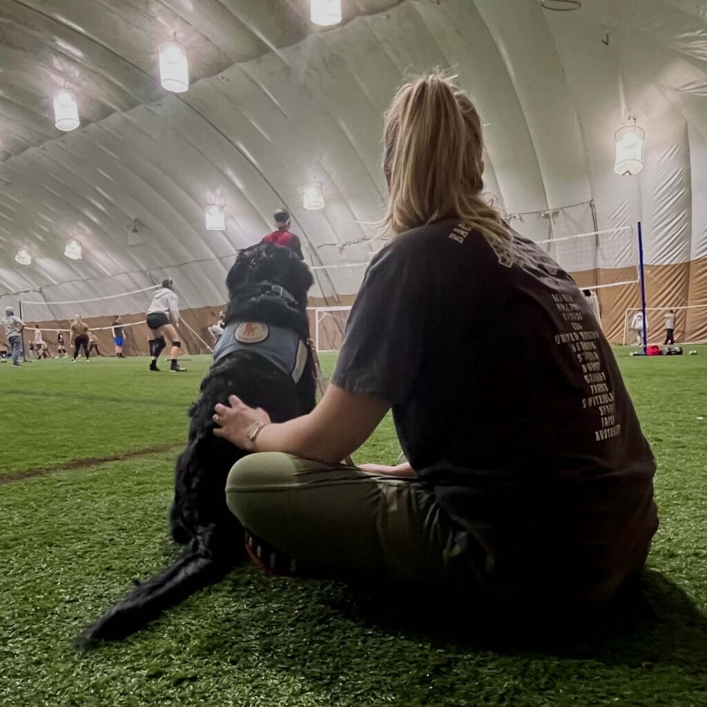 A person with a black Labrador/golden retriever dog in a training vest sits on the artificial turf of an indoor sports facility, observing a volleyball game. The spacious dome structure is well-lit with hanging lights, and players can be seen in action on the volleyball court.