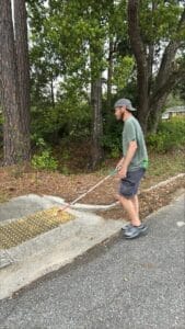 A young, bearded man wearing a backwards baseball cap, t-shirt and shorts, uses his cane to locate yellow, truncated domes at the edge of the sidewalk.