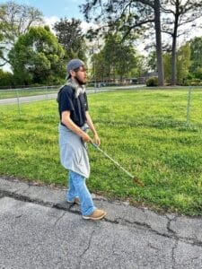 A young, bearded man wearing a backwards baseball cap, t-shirt and jeans, uses his cane to follow a grass line at the edge of the street.
