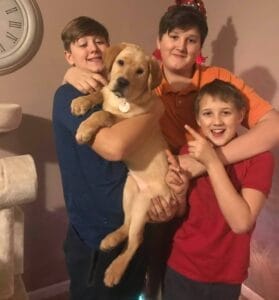 Three boys are hugging each other, while one of the boys is holding a yellow Labrador puppy. All boys and dog are look toward the camera smiling.