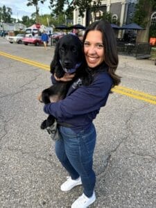 Uma pessoa com cabelo escuro de comprimento médio está em uma rua durante um evento de carro vintage. Ela está sorrindo em direção à câmera enquanto segura um jovem Labrador Retriever.
