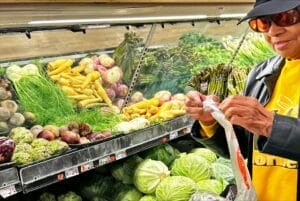 Uma pessoa fica na seção de produtos do supermercado, tentando abrir uma bolsa de produtos. Eles estão usando um chapéu escuro, camisa amarela e um casaco escuro.