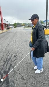 Uma pessoa usa uma bengala de suporte e uma longa bengala branca em uníssono enquanto atravessa um estacionamento. Eles usam óculos de sol, casaco escuro e jeans.