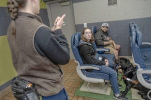 Uma mulher fica na frente de dois clientes que estão sentados em cadeiras de avião com seus cães a seus pés na instalação de treinamento do centro de Rochester. 