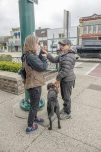 Duas mulheres usam ASL em uma esquina. O cliente tem um labrador preto no arnês no lado esquerdo e assina com a mão direita. 