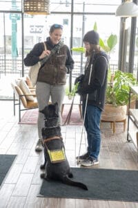 Duas mulheres ficam em um lobby brilhante cheio de janelas. Duas cadeiras e algumas plantas estão em segundo plano.