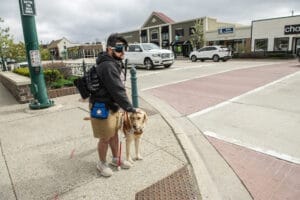Um homem está parado em um cruzamento de rua com um cão-guia preso. O homem está acariciando o topo da cabeça do cachorro enquanto segura uma longa bengala branca. O homem usa óculos de simulação de visão.