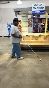 A man uses his white cane as he walks past a kiosk in the antique mall in which he runs a store. He is wearing a dark baseball cap, a gray t-shirt, and jeans.
