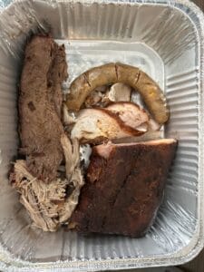 Five different types of smoked & barbecued meats arranged in a foil take-home tray