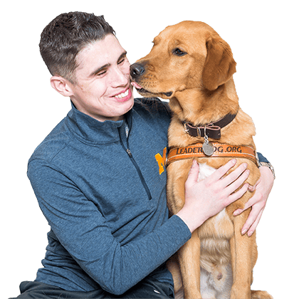 A young, brown-haired man smiles while hugging a reddish yellow lab/golden retriever cross. The dog is wearing a Leader Dog harness and licking the man's face.