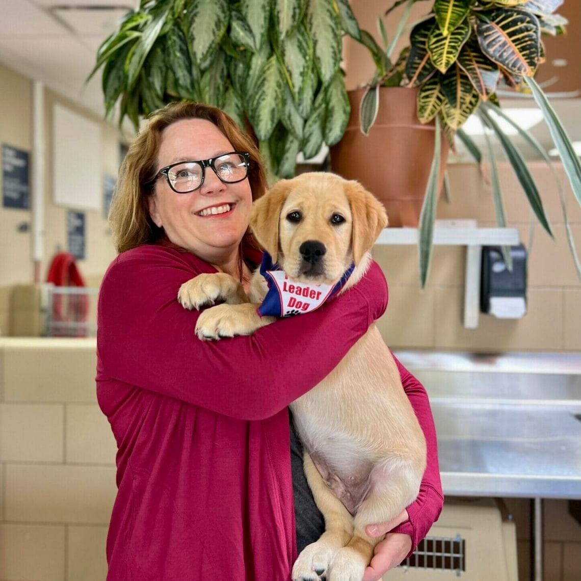 A woman in a maroon sweater smiles while holding a large yellow puppy wearing a future leader dog bandana