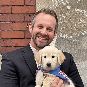 Dave Bann in a dark suit jacket and white shirt. He is smiling at the camera and holding a golden retriever puppy in its blue Future Leader Dog bandanna.