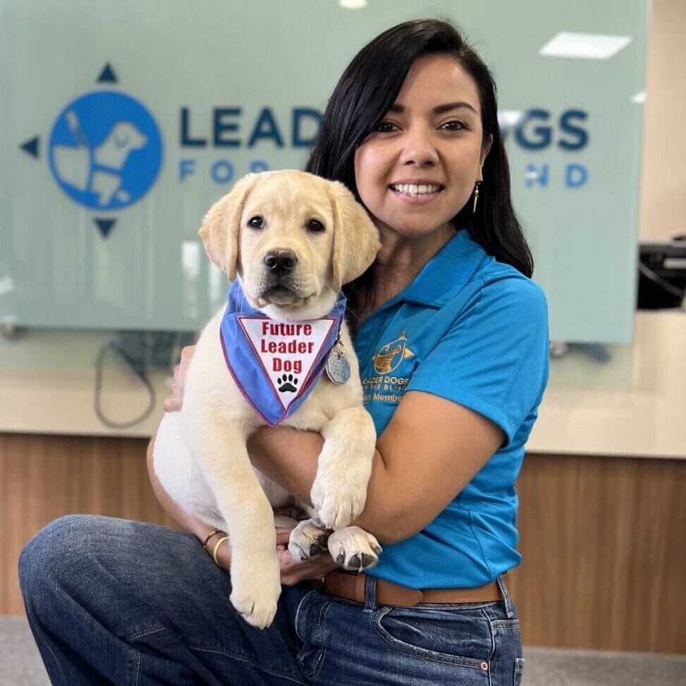 Gregory, Claudia with FLD Cowboy PREF A woman in a blue Leader Dogs for the Blind polo is kneeling in front of a Leader Dog sign while holding a yellow Labrador retriever puppy. The puppy is wearing a Future Leader Dog bandana.