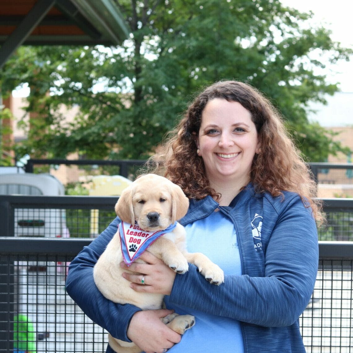 A woman in a blue Leader Dog sweater is outside holding a small yellow puppy with a future leader dog bandana