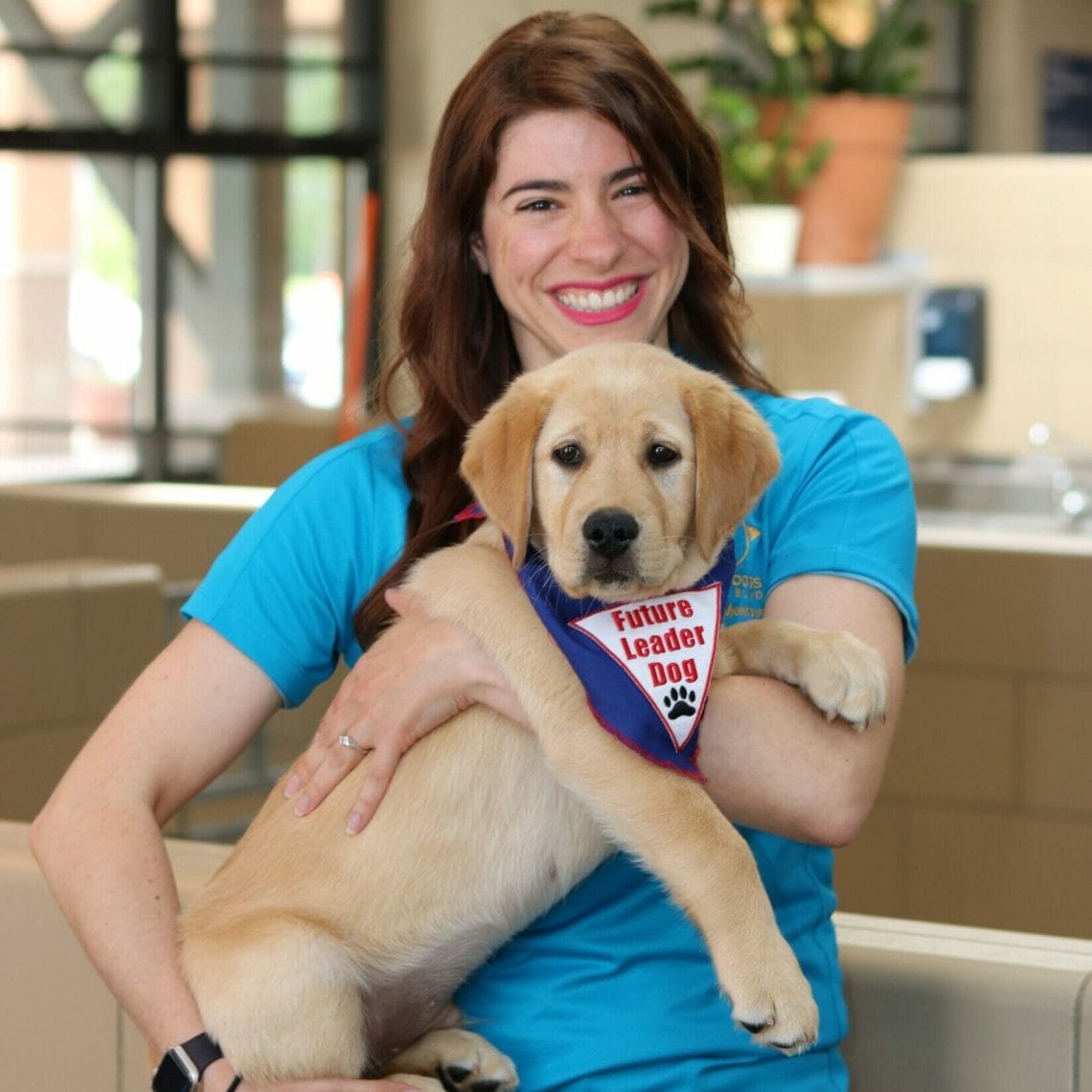 A woman with a big smile holding a yellow puppy with a Future Leader Dog bandana on