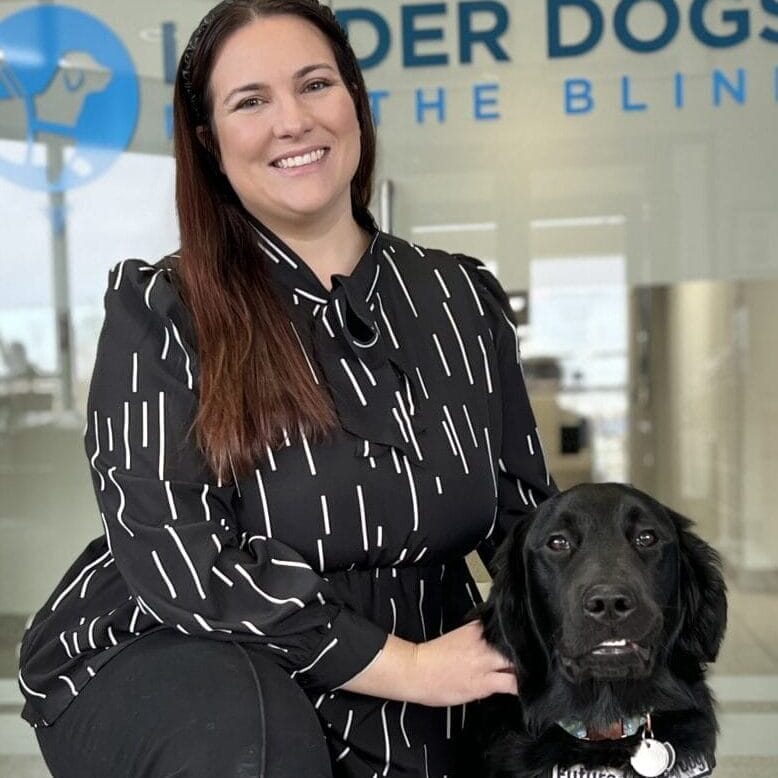 Martin Gardner_ Kelsey with Gram 2025 A woman with dark brown hair is kneeling next to a black Labrador/golden retriever Future Leader Dog. The are in front of a glass door with the Leader Dogs for the Blind logo on it.