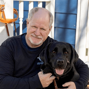 Tim sits outdoors on a sunny blue and white porch with his arms around a black lab that's sitting in front of him with its tongue out
