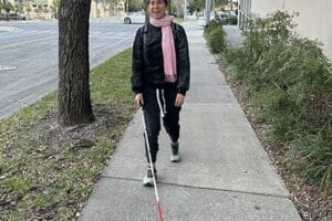 Smiling woman walking down a sidewalk toward the camera with a white cane. There is grass on either side of the sidewalk and some trees lining the sidewalk
